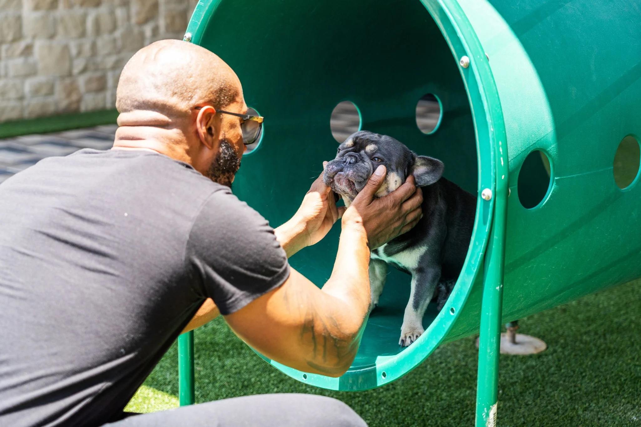 A man gently comforts a black-and-gray dog inside a bright green playground tunnel on artificial turf, creating a warm, caring moment.